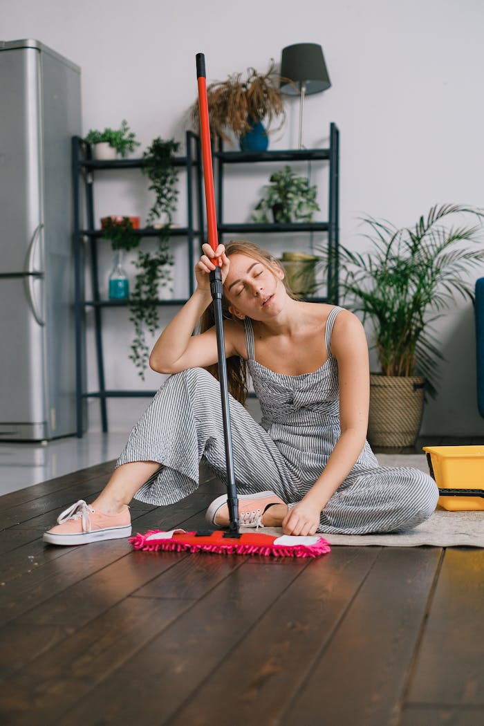 Exhausted young female with closed eyes leaning on handle of mop while sitting on carpet after washing floor in house